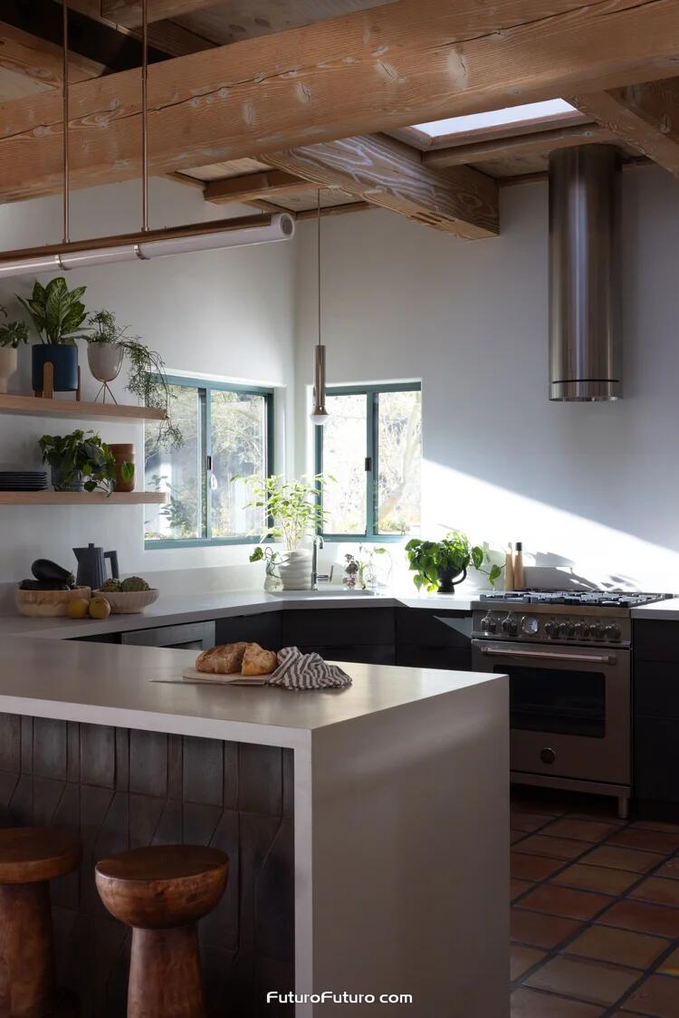 Polished stainless steel Futuro Futuro 14 inch Jupiter cylindrical wall range hood above a gas range in a Calabasas kitchen with wood beams, terracotta floors, white counters, and a dark island with stools.