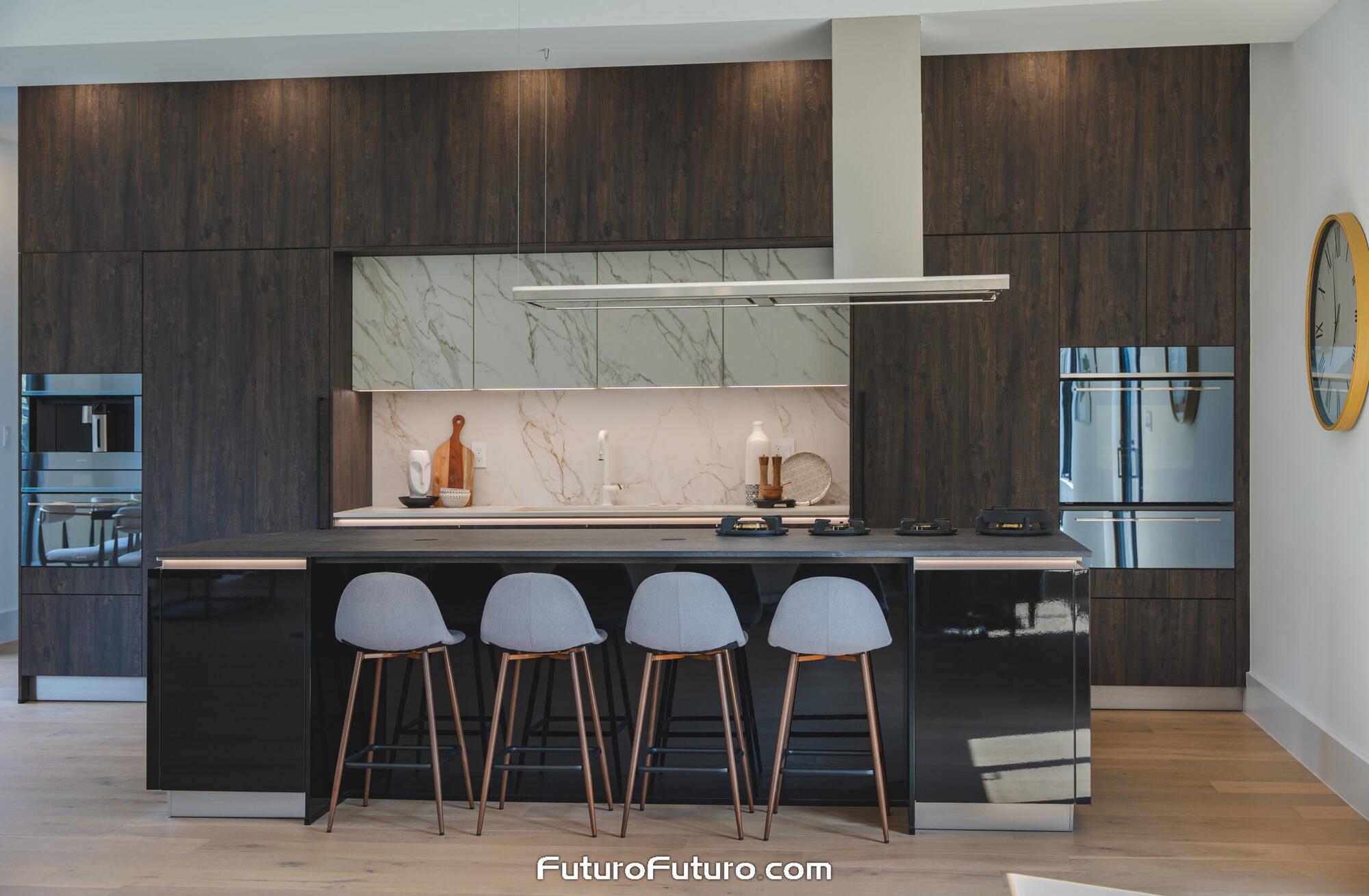 Slim stainless steel Futuro Futuro 69 inch Streamline island range hood suspended over a glossy black cooktop island with bar stools in a McLean Virginia kitchen of dark wood cabinets, marble niche, and ovens.