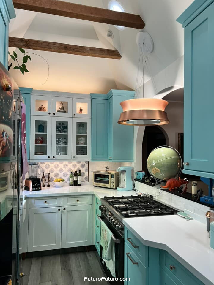 A copper Futuro Futuro 24 inch Arena island range hood hangs on thin mounting strings above a black gas range. Aqua cabinets, wood beams, a sloped white ceiling, patterned tile, and white counters fill this Altadena, California kitchen.