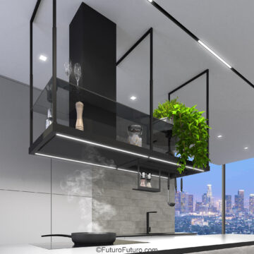 Side view of a matte black 69 inch island range hood over a white island, steam rising from a pan toward the canopy, glass shelves with wine glasses and a plant, hanging rack and utensils, grey tile and a dusk window.