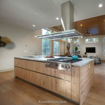 69 Inch Island Range Hood Open Vaulted Kitchen, Streamline Stainless Over White Island Gas Cooktop Luxury slim ceiling mount kitchen hood toward living room. Open kitchen with wood floors and a vaulted ceiling, slim stainless steel island range hood over a white veined island, LED strips lighting a teal kettle on a five burner gas cooktop, chimney to wood ceiling, sink at left, sofa beyond.